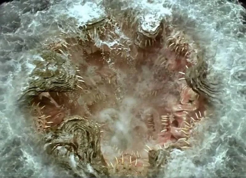 A swirling vortex in water revealing sharp, jagged formations resembling teeth, with frothy waves surrounding the opening.