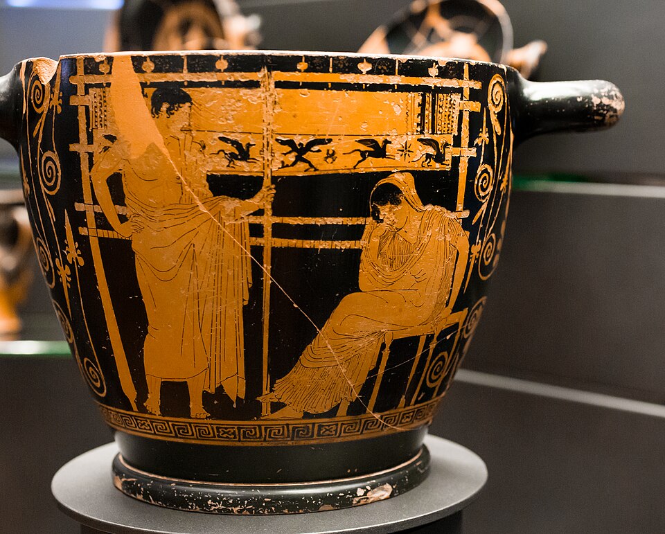 An ancient Greek vase with black and orange pottery, featuring a scene of two figures, one standing and the other seated, in classical attire against a decorative background.